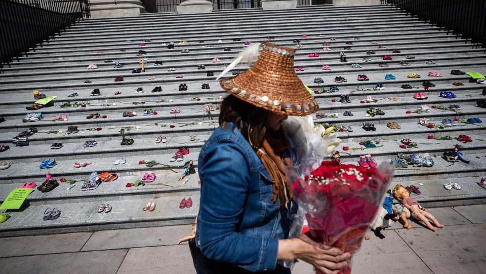 A woman carries flowers to be placed with 215 pairs of children's shoes on the steps of the Vancouver Art Gallery as a memorial to the 215 children whose remains have been found buried at the site of a former residential school in Kamloops, in Vancouver, on Friday, May 28, 2021. ( THE CANADIAN PRESS/Darryl Dyck)