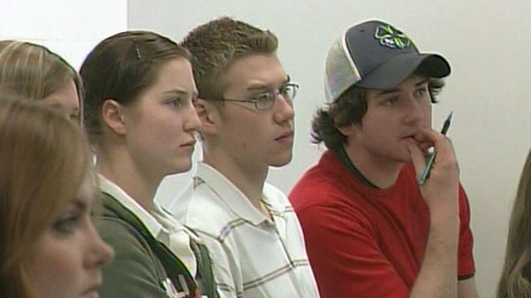 Students are seen in a classroom listening to a teacher. 