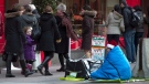 A homeless person sits on the sidewalk as holiday shoppers admire displays in the windows of a downtown department store in Toronto on Sunday, Dec. 23, 2012. (Frank Gunn / THE CANADIAN PRESS)