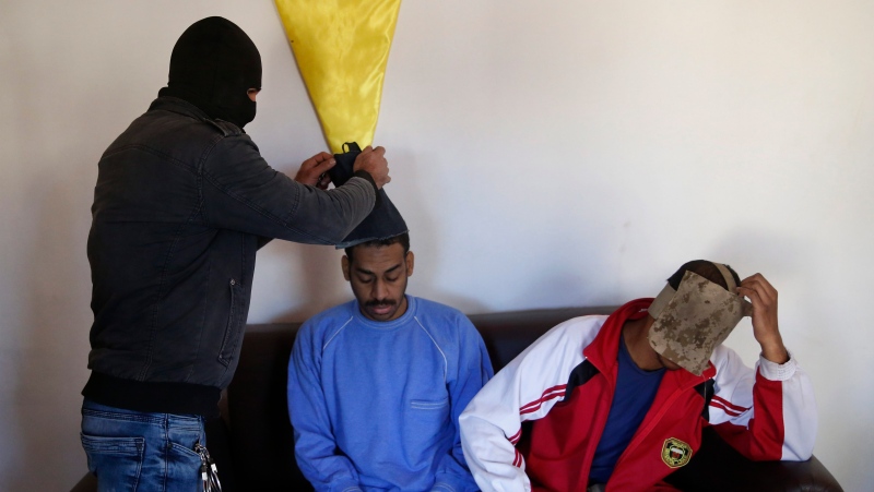 A Kurdish security officer takes off face masks from Alexanda Amon Kotey, left, and El Shafee Elsheikh, who were allegedly among four British jihadis who made up a brutal Islamic State cell dubbed "The Beatles," for an interview with The Associated Press at a security center in Kobani, Syria,  March 30, 2018. (AP Photo/Hussein Malla)