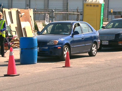 Cars waiting hours in line to drop off their garbage are blocked from driving into the waste transfer site by a large barrel.