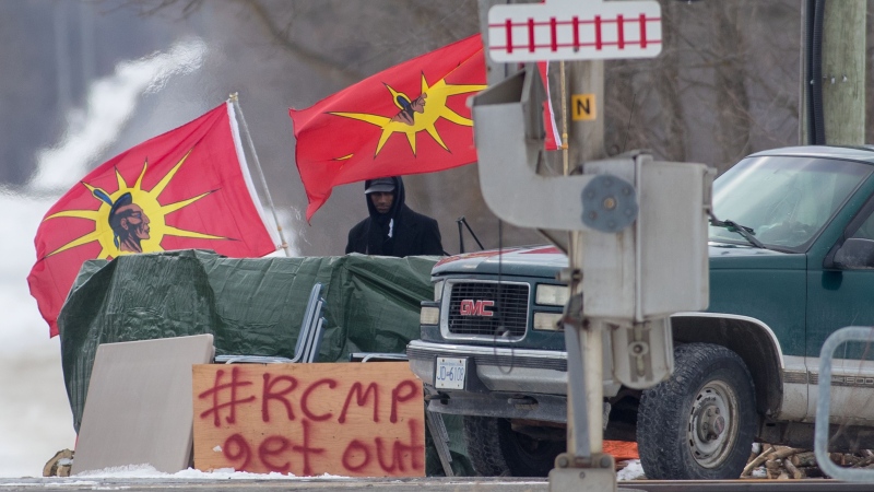 A protester stands between Mohawk Warrior Society flags at a rail blockade on the tenth day of demonstration in Tyendinaga, near Belleville, Ont., Sunday, Feb. 16, 2020. (THE CANADIAN PRESS / Lars Hagberg)