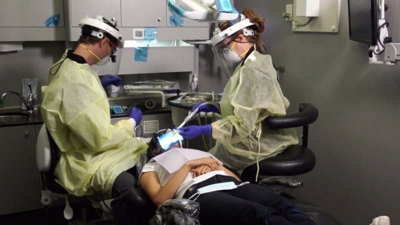 Two dentists in personal protective equipment (PPE) help a patient in this image. 