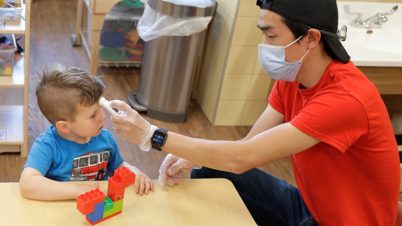 In this May 27, 2020 photo, a teacher at a daycare centre in Tacoma, Wash., wears a mask as he takes the temperature of a four-year-old boy. (AP Photo/Ted S. Warren)