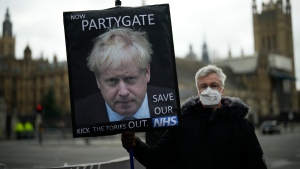 An anti-Conservative Party protester holds a placard with an image of British Prime Minister Boris Johnson including the words "Now Partygate" backdropped by the Houses of Parliament while in London on Dec. 8, 2021. (AP Photo/Matt Dunham)