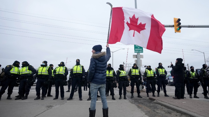 A supporter waves a Canadian flag on a hockey stick as police officers enforce an injunction against their demonstration, which has blocked traffic across the Ambassador Bridge by protesters against COVID-19 restrictions, in Windsor, Ont., Feb. 12, 2022. THE CANADIAN PRESS/Nathan Denette