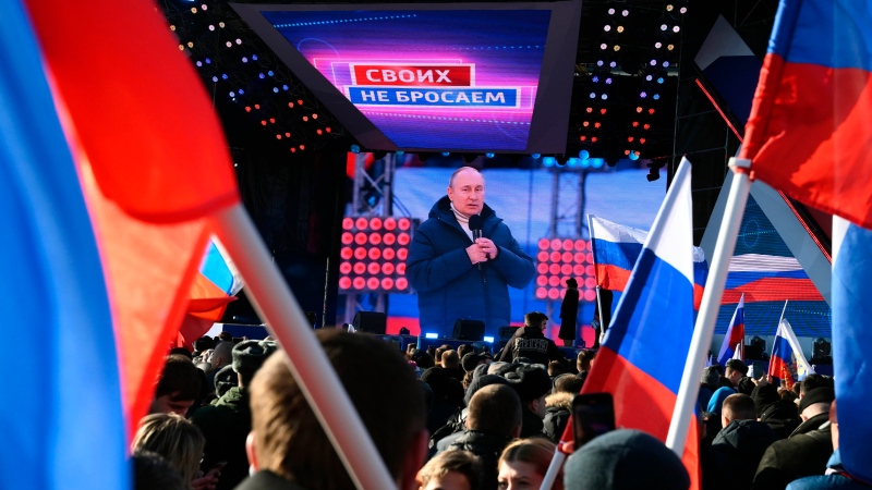 Russian President Vladimir Putin is seen on a big screen as he delivers his speech at the concert marking the eighth anniversary of the referendum on the state status of Crimea and Sevastopol and its reunification with Russia, in Moscow, Russia, Friday, March 18, 2022, with the words reading "We don't abandon our own" projected. (Sputnik Pool Photo via AP)