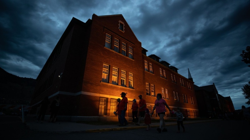 People are silhouetted as they walk past the former Kamloops Indian Residential School after gathering to honour the 215 children whose remains are believed to be buried near the facility, in Kamloops, B.C., on Monday, May 31, 2021. The year since the the Tk'emlups te Secwepemc First Nation announced that ground-penetrating radar had located the suspected grave sites in a former apple orchard has been one of national reckoning about residential schools in Canada. THE CANADIAN PRESS/Darryl Dyck