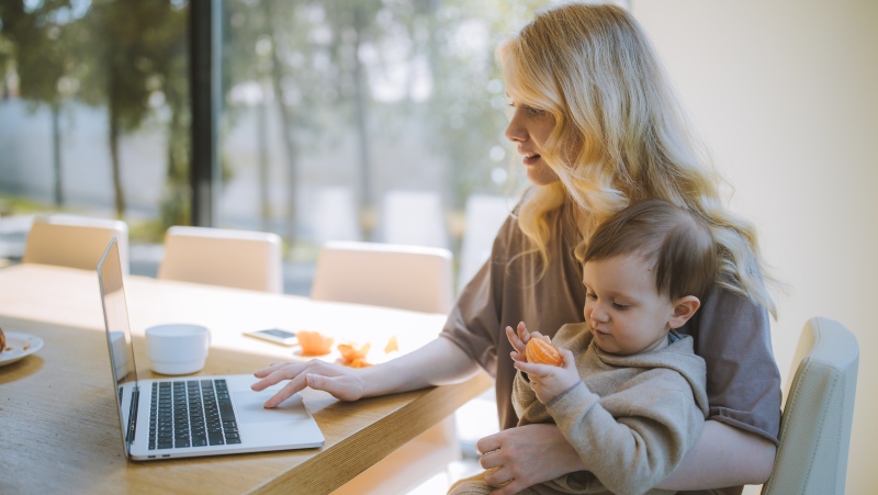 A woman is seen in this stock photo working on a laptop while holding a child. (Pexels)