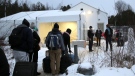 Migrants line up on the border of the United States, foreground, and Canada, background, at a reception center for irregular borders crossers, in Saint-Bernard-de-Lacolle, Quebec, Canada, Wednesday Jan. 12, 2022, in a photo taken from Champlain, N.Y. (AP Photo/Wilson Ring)