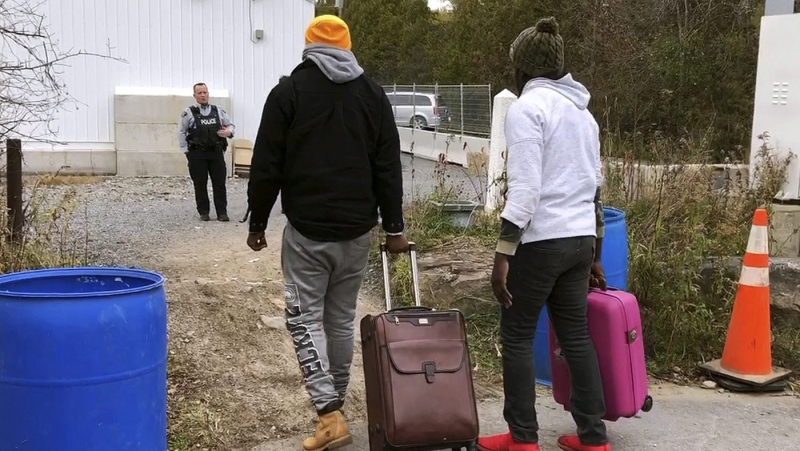 In this Nov. 4, 2019, photo, two men from Nigeria illegally cross the U.S. border at Roxham Road in Champlain, N.Y., into Canada where Royal Canadian Mounted Police stand, rear, in Saint-Bernard-de-Lacolle, Quebec. Since early 2017, when people who despaired of finding a permanent safe haven in the United States began turning to Canada for help, around 50,000 people have illegally entered Canada, many through Roxham Road in upstate New York. A case being heard in a Toronto court this week could end the use of Roxham Road. (AP Photo/Wilson Ring)