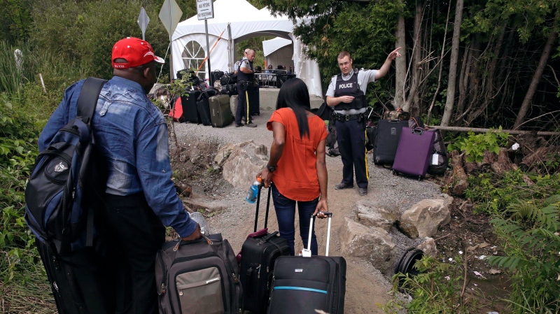In this Aug. 7, 2017 file photo, a Royal Canadian Mounted Police officer informs a migrant couple of the location of a legal border station, shortly before they crossed from Champlain, N.Y., to Saint-Bernard-de-Lacolle, Quebec, using Roxham Road. (AP Photo/Charles Krupa, File)