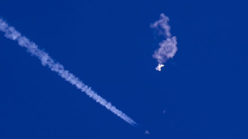 In this photo provided by Chad Fish, the remnants of a large balloon drift above the Atlantic Ocean, just off the coast of South Carolina, with a fighter jet and its contrail seen below it, on Feb. 4, 2023. The downing of the suspected Chinese spy balloon by a missile from an F-22 fighter jet created a spectacle over one of the state’s tourism hubs and drew crowds reacting with a mixture of bewildered gazing, distress and cheering. (Chad Fish via AP)