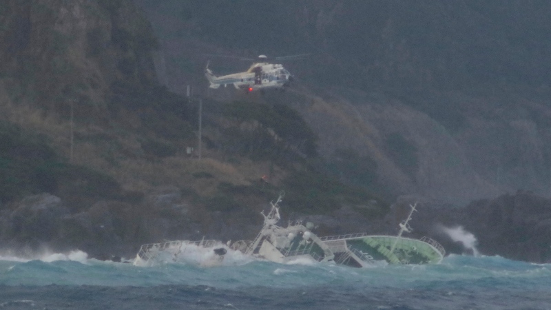 In this photo provided by Japan Coast Guard, a coast guard helicopter hovers over a fishing boat tilting almost to the side, for their rescue, off Kozushima, southwest of Tokyo, Monday, March 4, 2024. A tuna fishing boat went aground near one of Izu islands southwest of Tokyo after an engine trouble. ( Japan Coast Guard via AP)