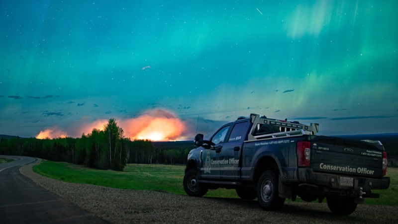 The Aurora Borealis shines overhead of a B.C. Conservation Officer Service vehicle near the junction of highways 97 and 77, as a wildfire burns in the background near Fort Nelson, B.C., in a Saturday, May 11, 2024, handout photo. THE CANADIAN PRESS/HO-Ministry of Water, Land and Resource Stewardship