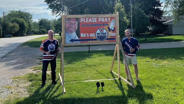 Mark Slater (left) and Helmut Reinhardt (right) pose in front of sign making fun of Slater's allegiance to the Oilers in Kingsville, Ont. on May 29, 2024.
