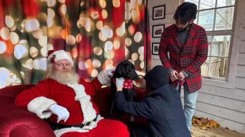 Santa Claus sits on a red couch next to a black dog while someone adjusts the dog's bandana.