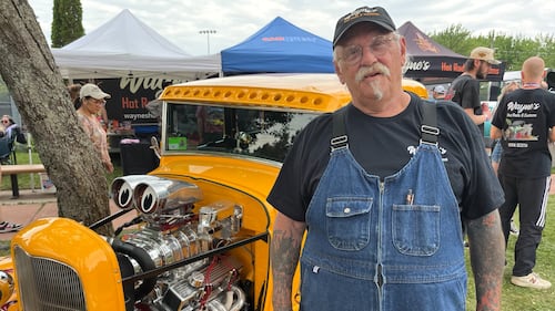 A man wearing denim overalls stands next to a yellow vintage car.
