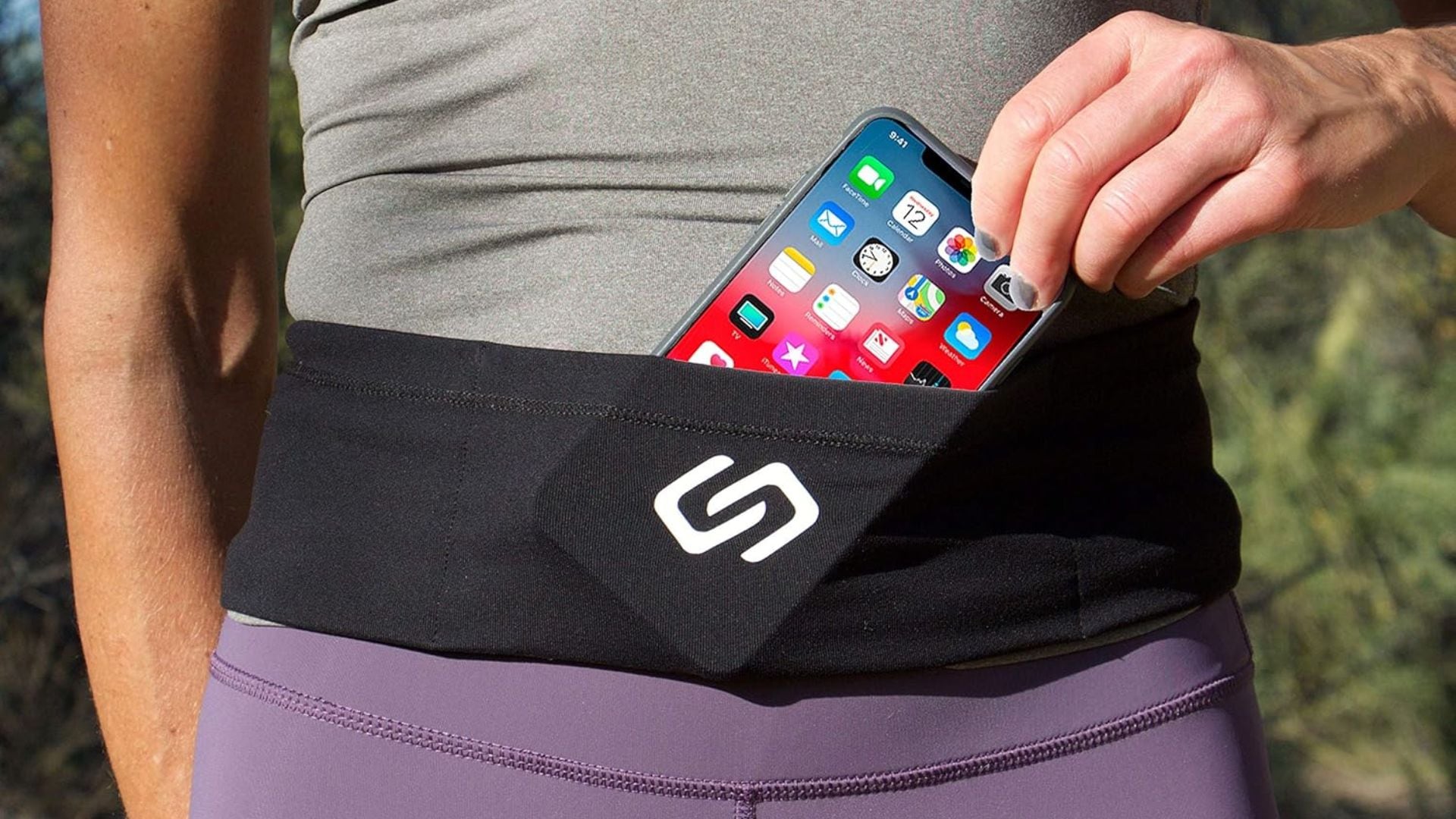 A person sliding their iPhone into their Sporteer running belt. They are wearing purple leggings and a grey tank top.