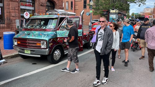 People walk in the street past classic cars.