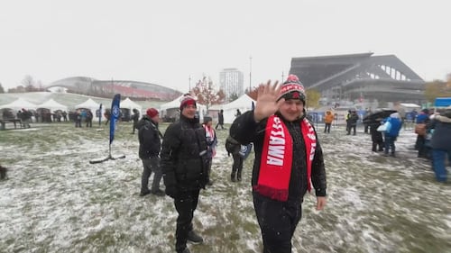 A Rapid FC fan wearing winter clothing waves at the camera surrounded by others dressed in similar fashion. They stand on the great lawn at TD Place, which is covered in snow.