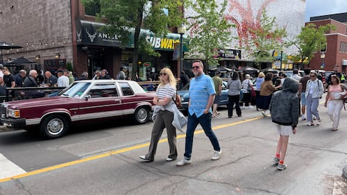 People walk in the street past classic cars.