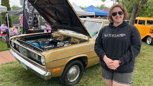 A woman in a black hoodie poses for a picture in front of a vintage car.